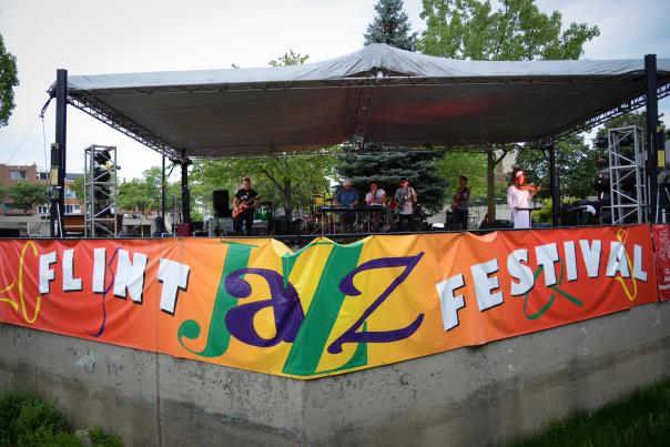 A picture of the front of the Riverbank Park stage during the Flint Jazz Festival with a band playing. There is a colorful banner on the cement stage reading FLINT JAZZ FESTIVAL.