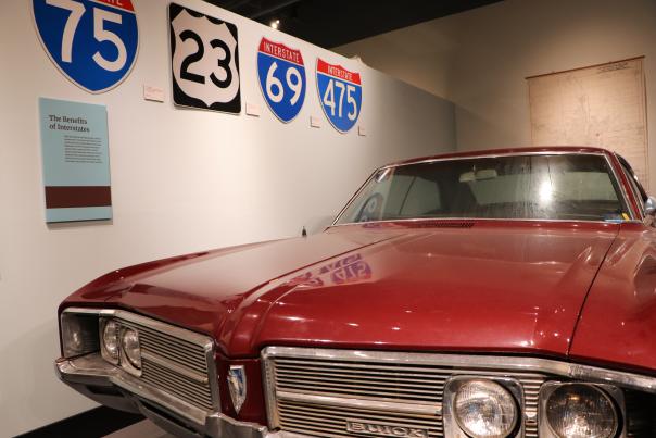 A red classic car on display at the Sloan Museum of Discovery with highway signs on the wall beside it.