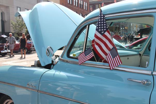 A blue classic car sits at a car show with American flags hanging out of its open window.