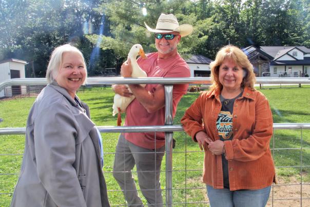 Two women pose outside on a farm while a man stands behind them holding a white duck.
