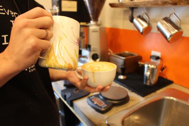 A person in a black t-shirt prepares a hot drink while creating an image in the foam on top of the drink.