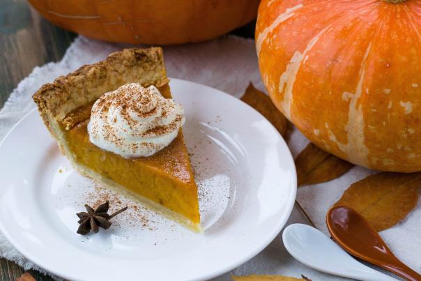 A slice of pumpkin pie on white plate beside two pumpkins on a table.