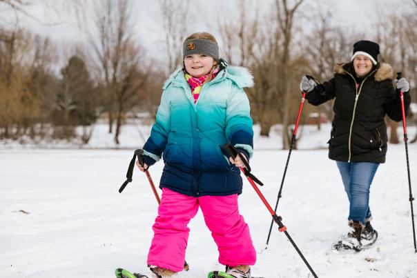 Young White girl dressed for winter and enjoying cross country skiing in the snow with a smiling woman skiing behind her.