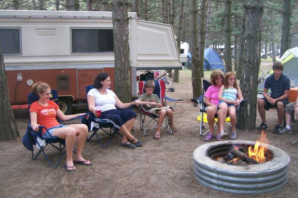 A family sits around a campfire at Wolverine Campground in Genesee County, Michigan with a brown and white pop-up camper in the background.