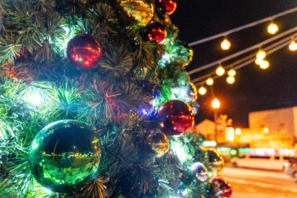 A close-up image of a Christmas tree lit up at night, with red and green ornaments reflecting the light in a downtown city setting.
