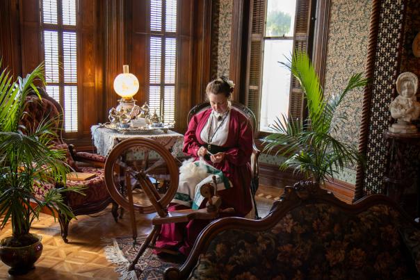 A woman wearing historic garb sitting before a spinning wheel at the Whaley Historic House Museum.