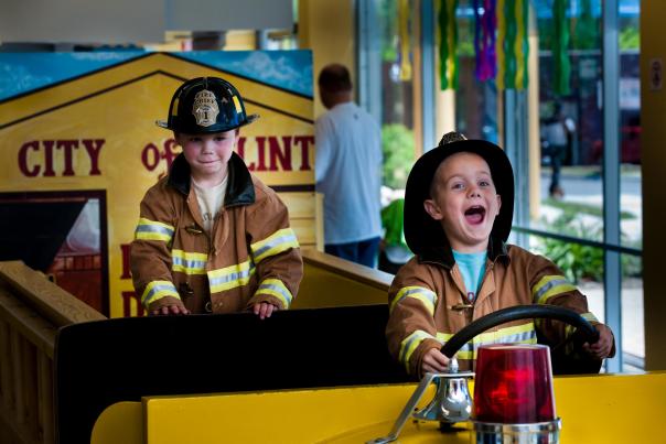 Two boys in fire fighter outfits, playing in a fake fire truck at the Flint Children's Museum.