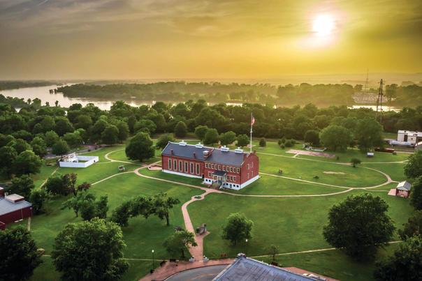 The sun is shining behind clouds above the National Historic Site in Fort Smith, Arkansas with the Arkansas River flowing behind it.