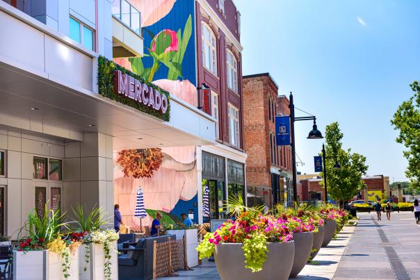 Street view of The Landing in Fort Wayne, Indiana with colorful murals, large potted flowers, and restaurants on a summer day