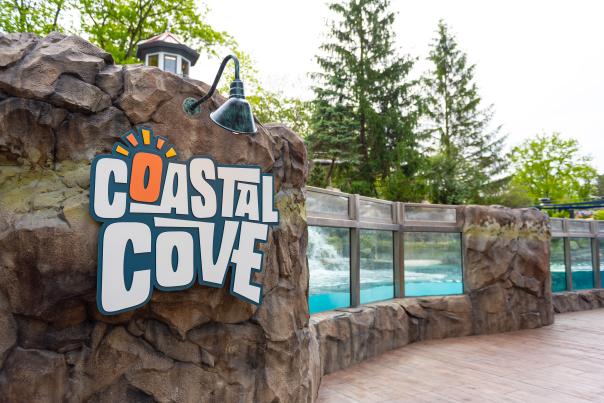 Close-up of the Coastal Cove exhibit sign at the Fort Wayne Zoo, with rock formations and underwater viewing windows for sea lions in the background.