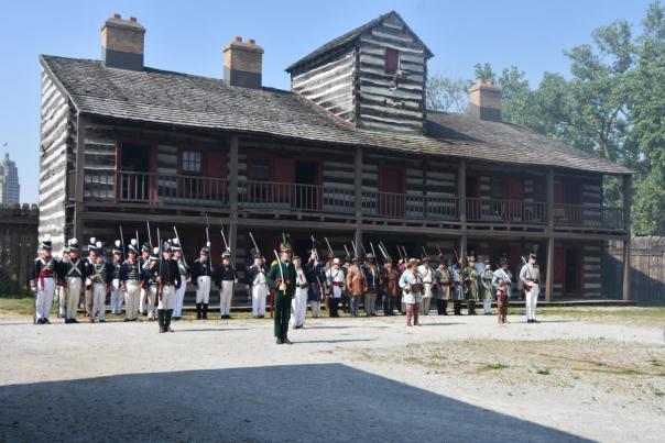 Re-enactors dressed as soldiers in uniform and holding historic weapons stand in formation in front of the Old Fort during a demonstration