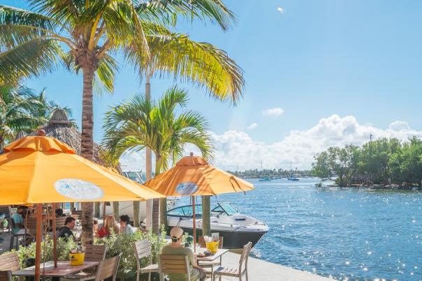 people dining along the Intracoastal Waterway under bright yellow umbrellas and palm trees on a beautiful, sunny day.