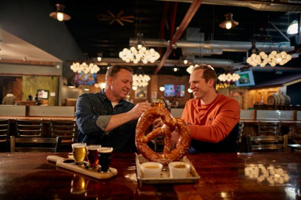 Two people sitting at a bar with a giant pretzel, a flight of beer, and they're cheers-ing one another.