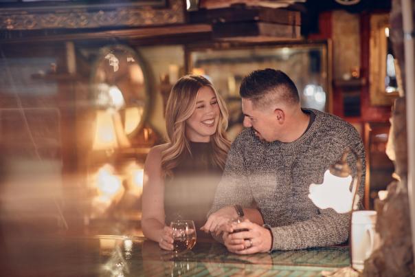 A couple standing at a bar, holding glasses of wine, and looking at eachother.