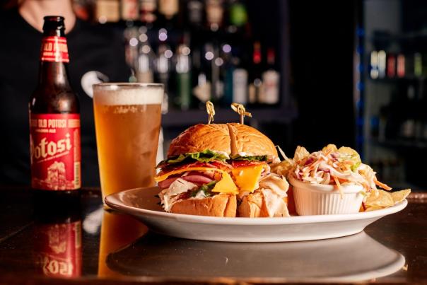 A plate of food with a sandwich, chips, coleslaw, and a pint of cold beer sitting on a bar.