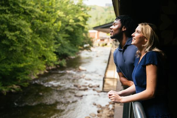 Guests standing on a hotel balcony overlooking a flowing mountain stream in Gatlinburg, Tennessee, surrounded by lush forest scenery and creekside lodging accommodations.