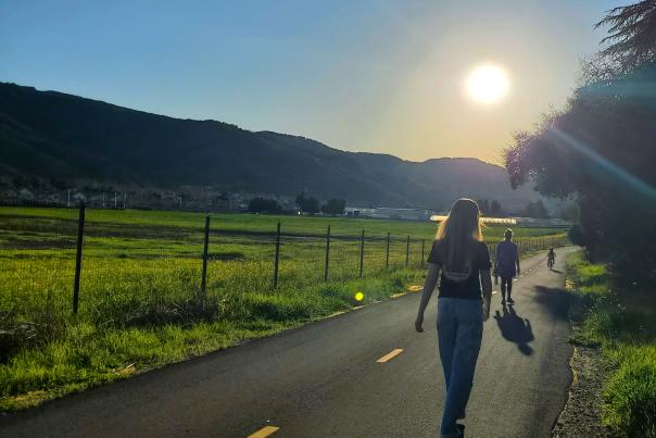 A paved walking and biking path with people walking and the sun shining, showing the surrounding greenery and mountain range