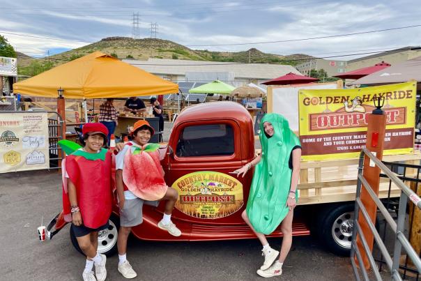 Three people in fruit and vegetable costumes posting by The Golden Hayride & Outpost Farmers Market truck and stands