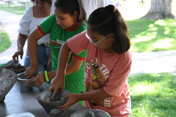Children learn to grind corn by hand on a metate, a bowl made from stone