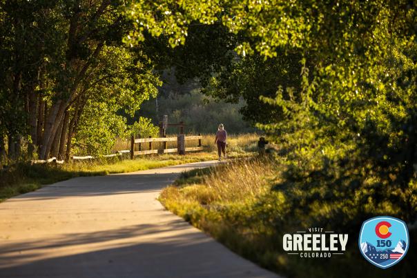 Trail in Greeley with woman walking far in the distance with Visit Greeley and 250-150 logos