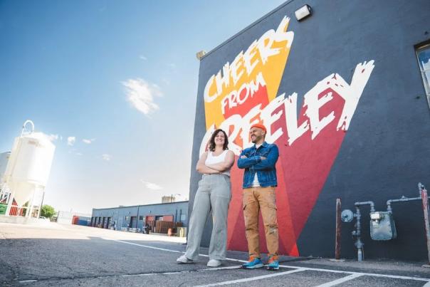 Armando and Briana smiling in front of the "Cheers from Greeley" mural.