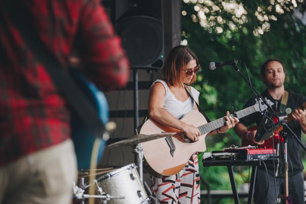 Woman wearing sunglasses playing a guitar with other band members nearby.