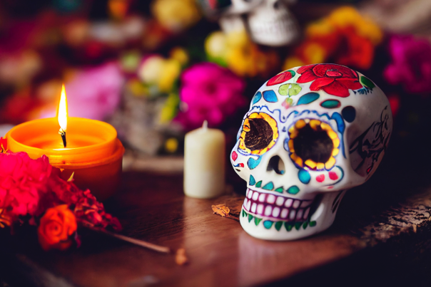 A small decorative skull with coloful design painted on it, next to a lit candle and some flowers