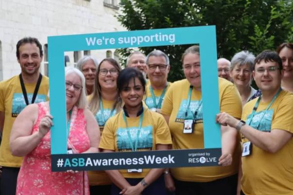 A group of people in yellow shirts and blue lanyards hold up a cardboard frame saying 'we're supporting ASB Awareness Week'