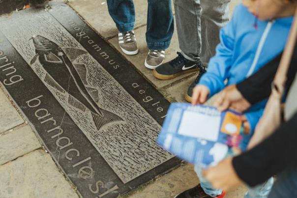 Stone plaque on Hull’s Fish Trail featuring an engraved fish illustration and species name, with visitors standing nearby while following the trail.