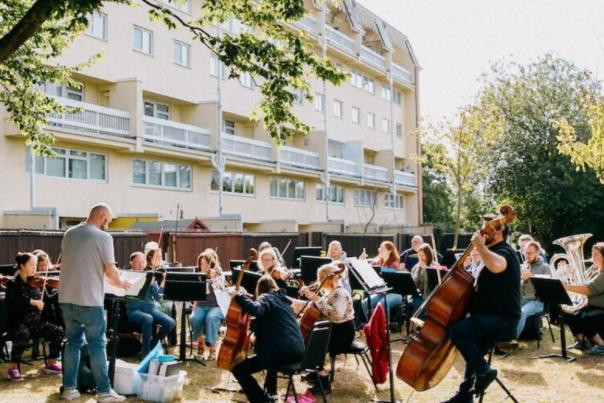 An orchestra plays outside near a block on flats on a sunny day