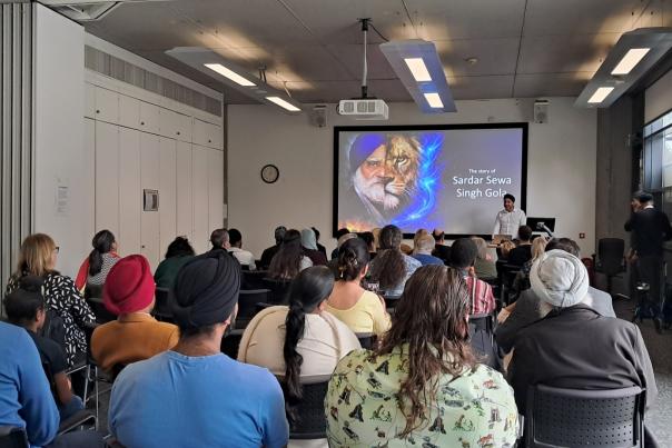 Attendees sit and watch Gulja Singh at the Hull History Centre talk about his father, an early Sikh settler in Hull. Gulja stands at a podium in a white shirt and black turban with a powerpoint slide which shows his father. Audience members sit on chairs looking ahead.
