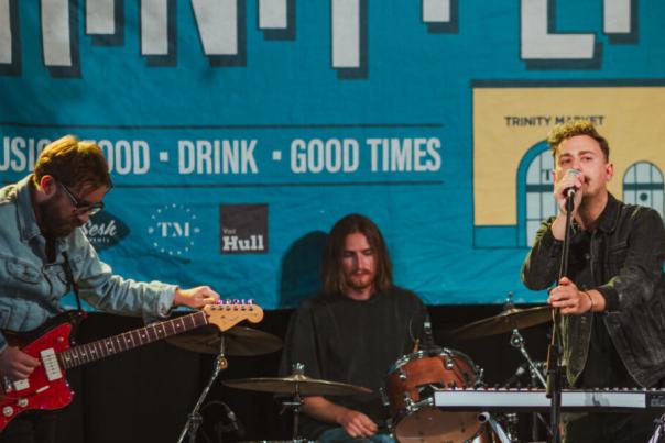 A band performs onstage in front of a blue banner that reads "LIVE MUSIC • FOOD • DRINK • GOOD TIMES." One member plays electric guitar, another plays drums, and the lead singer sings into a microphone while standing at a keyboard.