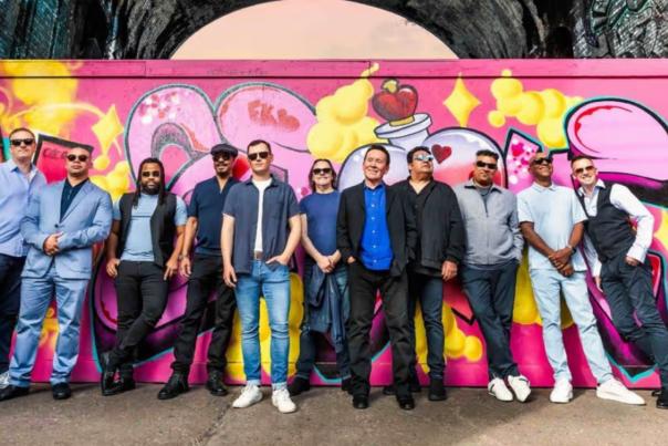 UB40 band stand in front of graffiti art. 11 men, some in sunglasses lean against the graffiti art which is sprayed with bright pink and red paint.