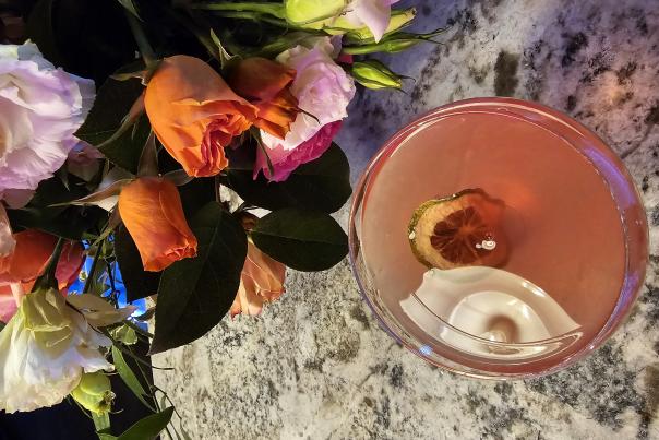 A pink cocktail garnished with a dried citrus slice sits on a marble bar beside a colorful bouquet of roses and fresh flowers at The Dirty Olive in Huntsville, Alabama. The close-up view and warm lighting create an elegant and upscale cocktail lounge atmosphere.