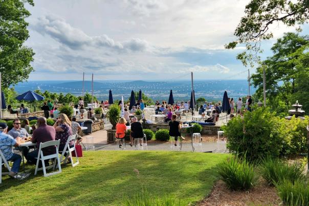 Guests gather on the scenic terrace at Burritt on the Mountain, enjoying food, drinks, and conversation while overlooking panoramic views of Huntsville and the Tennessee Valley. White tables, string lights, and lush greenery surround the outdoor space, creating a lively atmosphere against a backdrop of rolling mountains and dramatic summer skies.