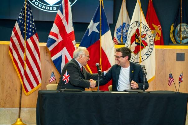 Mayor Watson of Austin and Mayor Burnham of Manchester sit behind desk and shake hands in front of national flags at the City Hall