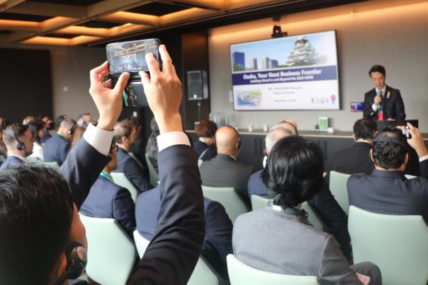 An audience member uses his phone to take a photo of Mayor Yokoyama of Osaka, Japan presenting to a business audience wearing suits at the Building Bridges Breakfast Seminar in Manchester during Japan Week 2025.