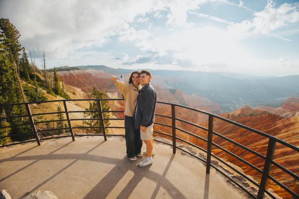 A couple stands at a paved scenic overlook at sunset, taking a selfie with the bright orange and red rock formations of Cedar Breaks National Monument behind them.