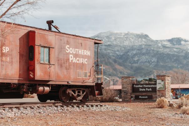 A weathered orange caboose on a small spur of track with the words SOUTHERN PACIFIC painted in white on the side sits in front of a snowy mountain landscape with a rock sign reading "Frontier Homestead State Park Museum" in front of it.