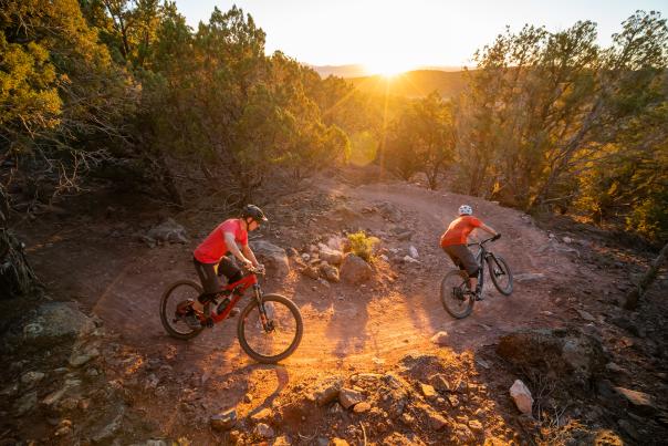 Two cyclists rounding a turn on the Lichen It mountain bike trail in Cedar City Utah at sunset with a sunburst over the distant hills.
