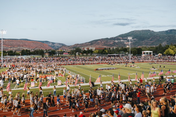 Large crowd gathered at a Cedar City football stadium with American flags and red rock mountains in the background at sunset