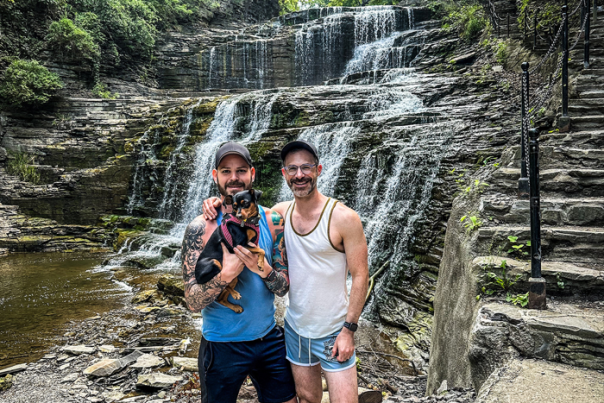 Two men stand in front of a waterfall, holding a small dog.