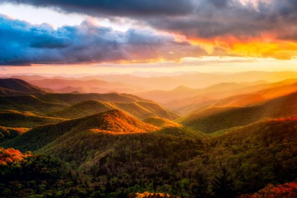 A dramatic sunset over the rolling peaks of the Blue Ridge Parkway, with intense golden light illuminating the mountain ridges beneath a colorful, cloudy sky.