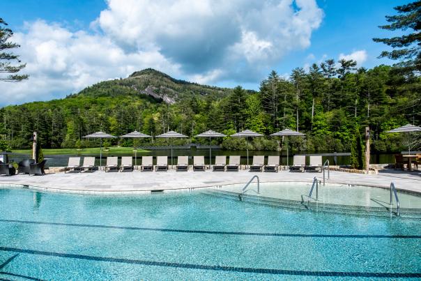 A scenic, wide-angle photograph of a luxury outdoor swimming pool with light blue water and black lane lines. The paved stone deck features a long, neat row of approximately nine empty beige sun loungers, each positioned under an open black-and-white striped umbrella. A rustic fence separates the deck from a calm lake. Beyond the lake, a prominent, forested mountain with large exposed rock faces rises dramatically under a bright blue sky filled with puffy white cumulus clouds, framed by tall pine trees. The scene is sun-drenched and peaceful.