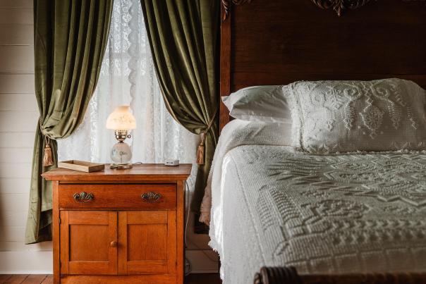 A close-up of a vintage-style bedroom. An antique wooden nightstand with a small floral glass lamp sits beside a bed featuring a high, ornately carved wooden headboard and a white textured bedspread. Thick green velvet curtains with gold tassels frame a window in the background.