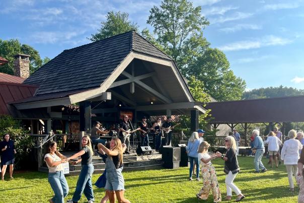 A band plays live music on a rustic wooden stage under a pavilion. Multiple groups of people, including teenagers on the left wearing denim and adults on the right wearing casual attire, dance enthusiastically on a bright green grassy lawn on a sunny day. Trees frame the background.