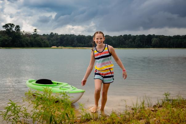 A little girl pulls a kayak out of the lake while smiling at the camera