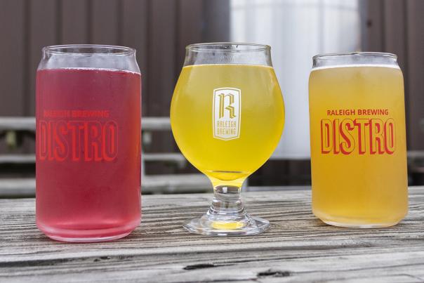 Three glasses of various alcoholic beverages sit on a wooden picnic table outside of Raleigh Brewing Distro in Smithfield, NC.