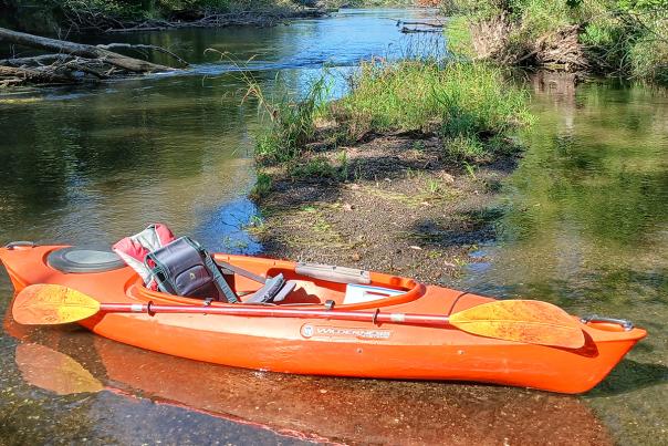 A bright orange kayak rests half in the water and half on a rocky shore in the Kalamazoo River.