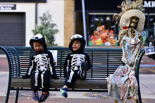 2 kids in skeleton costumes sitting on a bench next to a dressed up skeleton